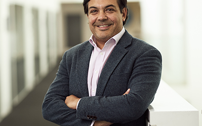 A man wearing a suit stands smiling. He is crossing his arms. He is standing in a hallway.