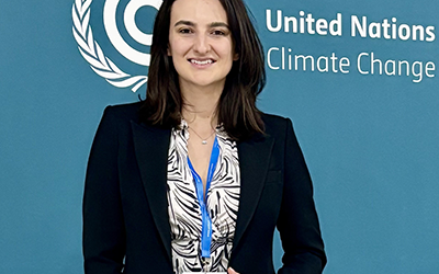 A woman with shoulder-length brown hair smiles. She is wearing a dark suit, a patterned shirt and a lanyard. She is standing in front of a backdrop that reads "United Nations Climate Change" on it.