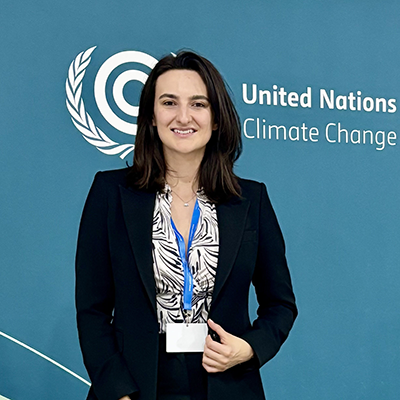 A woman with shoulder-length brown hair smiles. She is wearing a dark suit, a patterned shirt and a lanyard. She is standing in front of a backdrop that reads "United Nations Climate Change" on it.