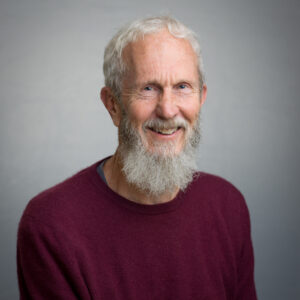 A man with a white hair and full white beard smiles at the camera. He is wearing a dark-coloured jumper.