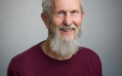 A man with a white hair and full white beard smiles at the camera. He is wearing a dark-coloured jumper.