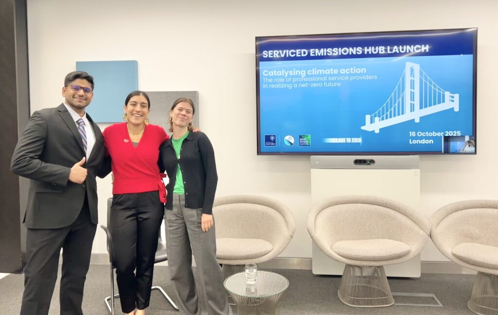 A man and two women stand at the front of a room with their arms around each other. The man is wearing a suit giving a thumbs up. Behind them are chairs and a screen that has a presentation on it.