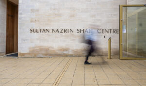 A person walks in front of a building. They are mid-stride. They appear blurry and in motion while the background is in focus. The name of the building is visible and says "Sultan Nazrin Shah Centre".