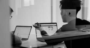 Two people sit at a table in an office. They are seen through a window. One of them is gesturing. They are sitting in front of laptops. Other laptops are reflected on the window.