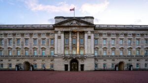 A photo of Buckingham Palace from the front. The building is made of stone and has many windows and a long balcony. There is a UK flag flying from the top.