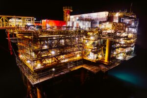 A large oil rig stands in the water. It is nighttime and the rig is lit up brightly.