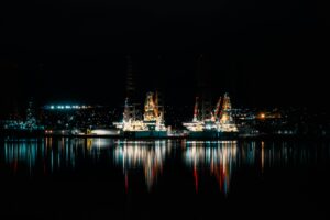 Two large oil platforms on the water. It is nighttime and they are lit up. The lights of a city are behind them.