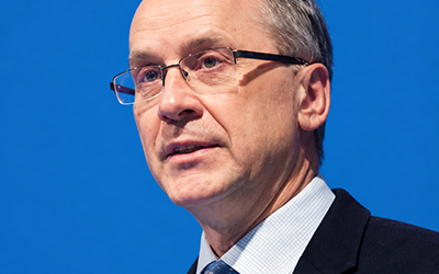 A man wearing a blazer, tie and collared shirt gazes ahead as he speaks. He is turned slightly to the side. He has short hair and is wearing glasses.