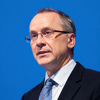 Brendan Devlin portrait_cropped A man wearing a blazer, tie and collared shirt gazes ahead as he speaks. He is turned slightly to the side. He has short hair and is wearing glasses.