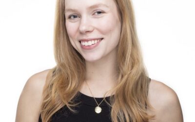A woman with long hair smiles. She wears a sleeveless top and a pendant necklace.